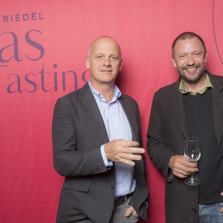 Zwei Männer in Anzügen lächeln vor einem rosa Hintergrund mit dem Text „Riedel Glas Tasting“. Einer hält ein Weinglas, der andere gestikuliert mit der Hand.