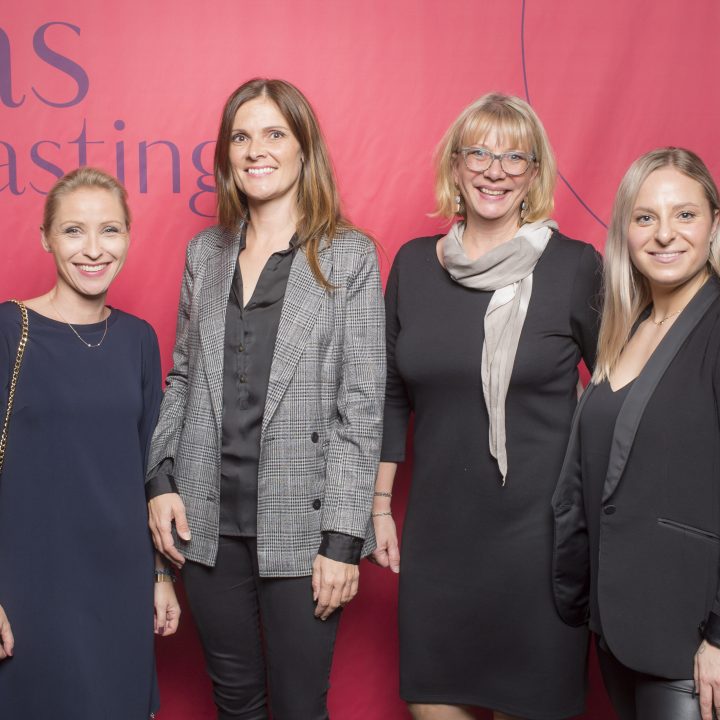 Vier Frauen stehen vor einem roten Hintergrund mit der Aufschrift „Glas Tasting“. Sie lächeln und sind unterschiedlich gekleidet, von leger bis Business-Kleidung.