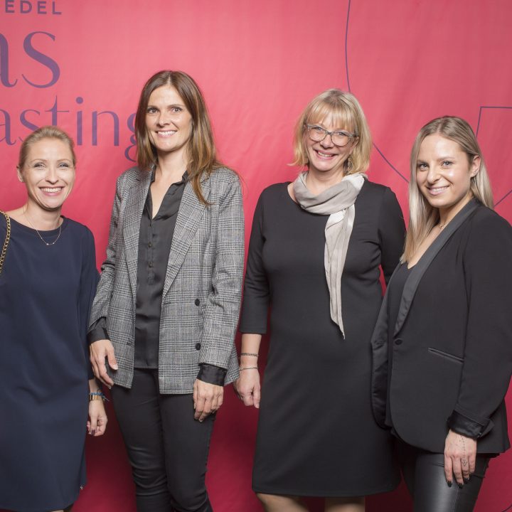 Vier Frauen stehen vor einem roten Hintergrund mit der Aufschrift „Riedel Glas Tasting“. Sie sind in formelle Kleidung gekleidet, lächeln und posieren für das Foto.