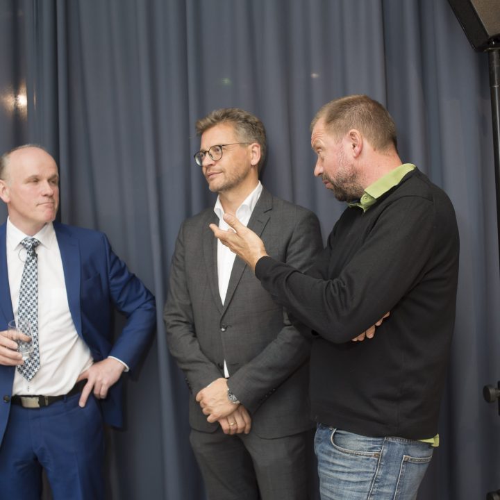Drei Männer stehen vor einem dunklen Vorhang und unterhalten sich. Der Mann links trägt einen blauen Anzug, der Mann in der Mitte einen grauen Anzug und der Mann rechts trägt einen schwarzen Pullover und Jeans. Sie scheinen in ein Gespräch vertieft zu sein.