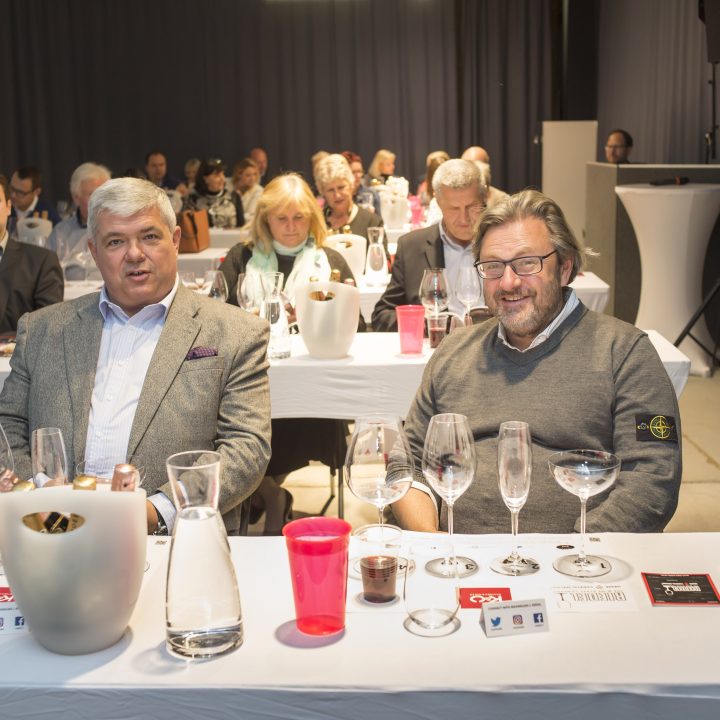 Eine Gruppe von Menschen sitzt an Tischen mit weißen Tischdecken, auf denen Weingläser und Krüge stehen. Zwei Männer sitzen lächelnd im Vordergrund. Andere Teilnehmer sitzen in Reihen hinter ihnen und konzentrieren sich auf eine Präsentation.