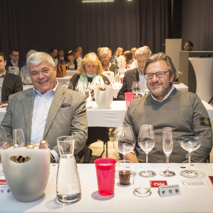 Eine Gruppe von Menschen sitzt an Tischen in einer Konferenz- oder Seminarumgebung. Zwei Männer stehen im Vordergrund und lächeln mit Gläsern und Flaschen auf dem Tisch. Andere im Hintergrund blicken nach vorne, hinter ihnen befindet sich ein grauer Vorhang.