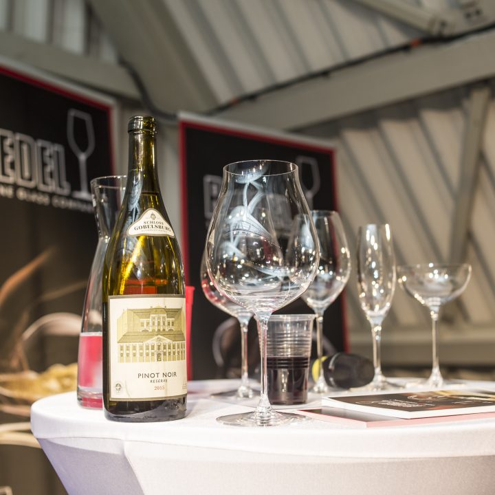 Auf einem runden Tisch mit weißer Tischdecke stehen eine Flasche Pinot Noir, mehrere Weingläser und eine Broschüre. Im Hintergrund sind Markenbanner mit Weinglasbildern zu sehen.