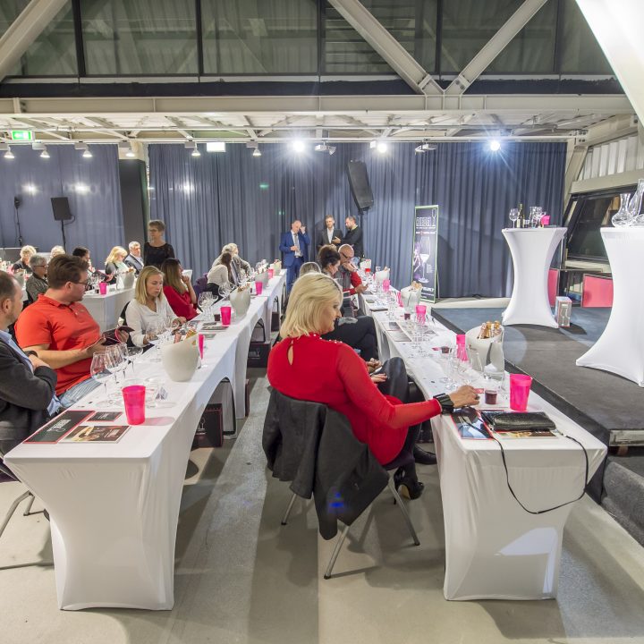 Eine Gruppe von Menschen sitzt an Tischen in einem modernen Innenbereich mit hohen Decken und freiliegenden Balken. Die Teilnehmer unterhalten sich. Auf den Tischen liegen Gläser und Papiere, was wahrscheinlich auf ein Seminar oder eine Verkostungsveranstaltung hindeutet.