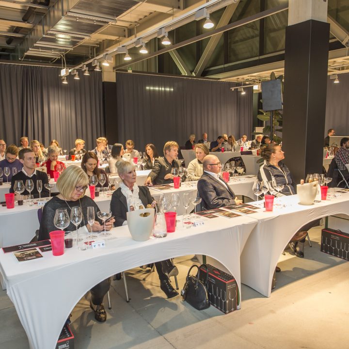 Eine Gruppe von Menschen sitzt an Tischen, die wie ein Klassenzimmer angeordnet sind. Jeder hat Weingläser, Wasserflaschen und rote Tassen dabei. Der Raum ist gut beleuchtet, mit grauen Vorhängen und Deckenlampen. Die Teilnehmer scheinen einer Präsentation oder Vorlesung aufmerksam zuzuhören.