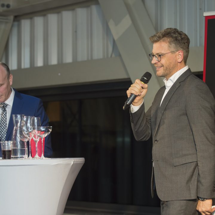 Zwei Männer in Anzügen stehen mit Weingläsern an Stehtischen. Einer spricht in ein Mikrofon, während der andere eine Weinflasche untersucht. Im Hintergrund ist teilweise ein Banner mit dem Wort „RIEDEL“ zu sehen.
