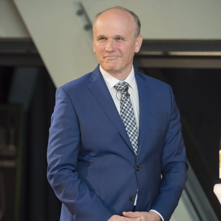 Ein Mann in einem blauen Anzug und einer gemusterten Krawatte steht vor einem grauen Industriehintergrund, lächelt leicht und hat die Hände gefaltet.
