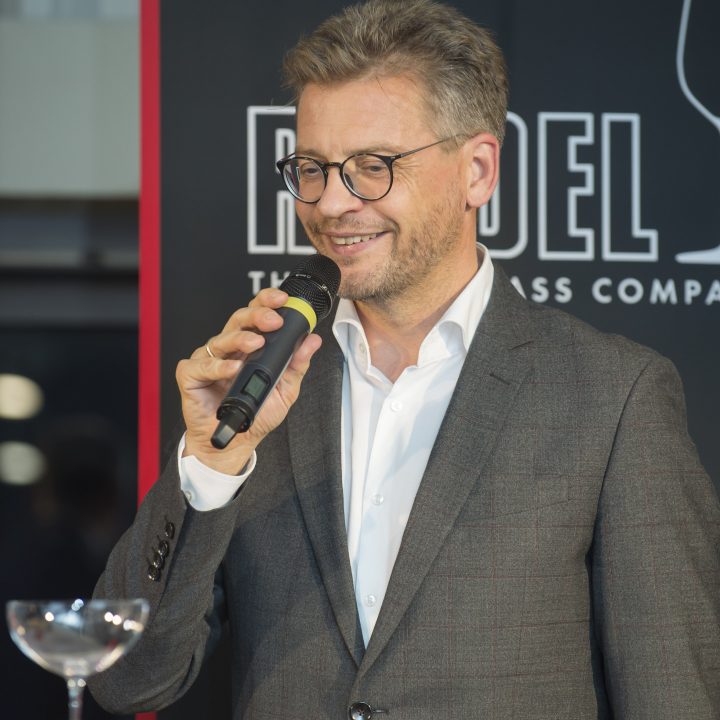 Ein Mann in grauem Anzug und Brille spricht in ein Mikrofon. Er steht neben einem Tisch, auf dem ein Weinglas steht. Im Hintergrund ist ein schwarz-rotes Banner mit teilweise sichtbarem Text zu sehen.