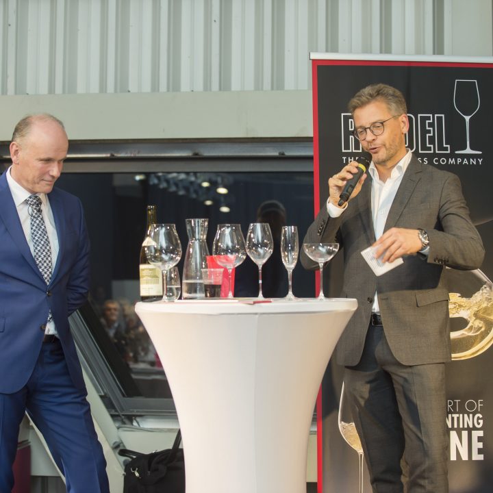 Zwei Männer in Anzügen stehen an einem runden Tisch mit Weingläsern und einer Flasche. Einer hält ein Mikrofon und spricht, während der andere zuhört. Sie sind drinnen, im Hintergrund sind Banner mit dem Wort „Riedel“ zu sehen.