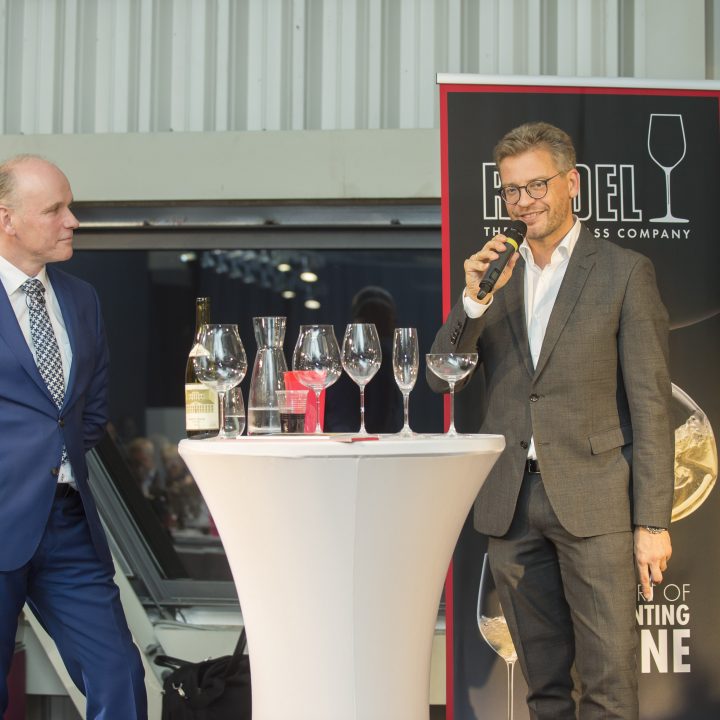 Zwei Männer in Anzügen stehen mit Weingläsern und einer Flasche an einem hohen Tisch. Ein Mann hält ein Mikrofon und spricht, während der andere zuhört. Sie befinden sich in einem modernen Veranstaltungsort mit Metallwänden und großen Fenstern. Im Hintergrund ist ein Markenlogo zu sehen.