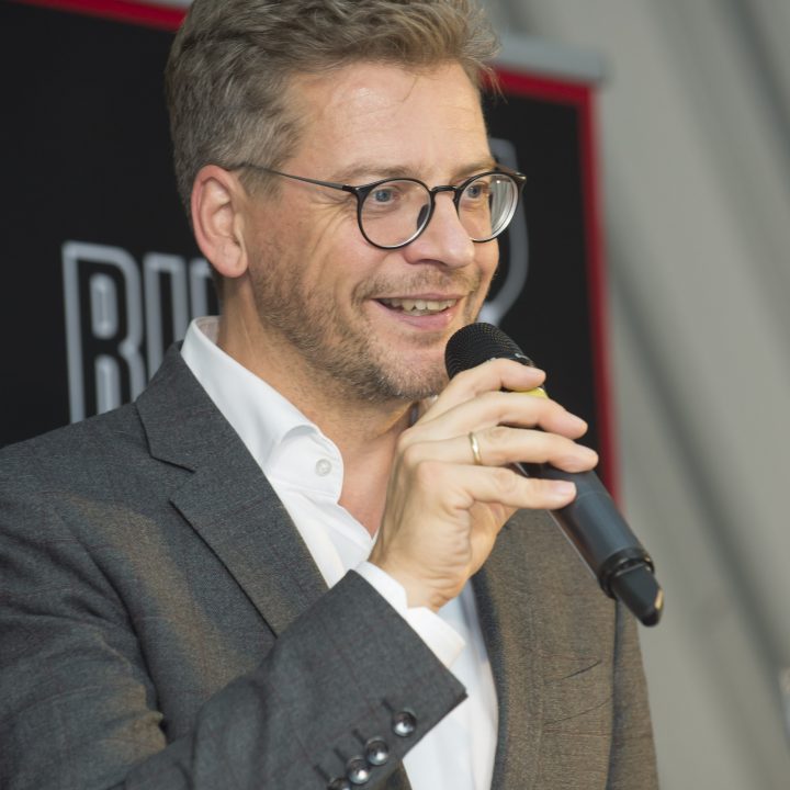 Ein Mann in grauem Anzug und Brille spricht in ein Mikrofon. Er hat kurze Haare und lächelt. Im Hintergrund ist ein teilweise sichtbares Logo in den Farben Rot und Schwarz zu sehen.