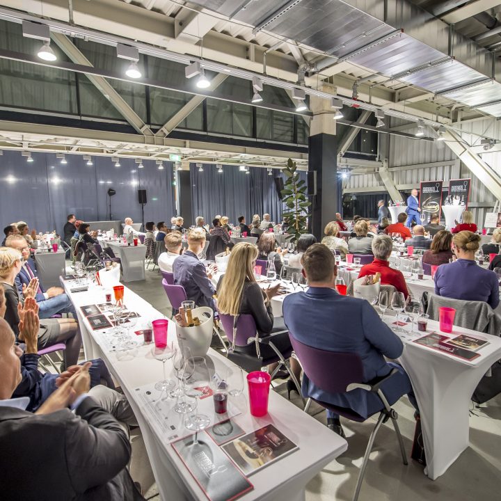 Eine große Gruppe von Menschen sitzt an Tischen in einem Konferenzraum. Der Raum ist gut beleuchtet und modern eingerichtet. Die Teilnehmer sind formell gekleidet, einige diskutieren. Präsentationsmaterialien und Getränke stehen auf den Tischen.