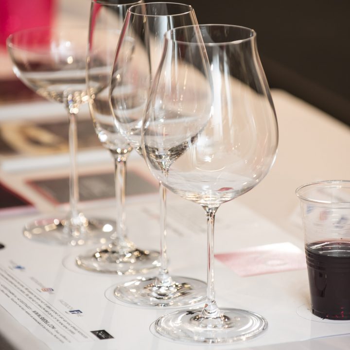 Auf einem Tisch steht eine Reihe leerer Weingläser mit Informationsblättern, daneben ein durchsichtiger Plastikbecher mit Rotwein.