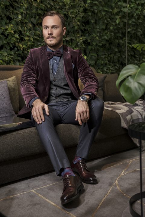 Ein Mann in Abendgarderobe, bekleidet mit einem burgunderfarbenen Samtjackett, einer Fliege und einer grauen Hose, sitzt auf einem Sofa vor einer grünen Wand und blickt zur Seite.