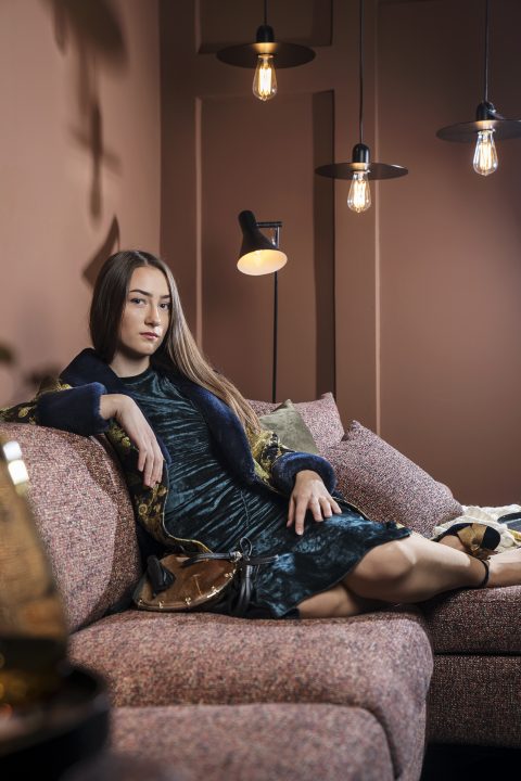 Eine Frau mit langen Haaren entspannt sich auf einem strukturierten Sofa in einem warm beleuchteten Raum. Sie trägt ein dunkelgrünes Kleid und eine gemusterte Jacke, ihr Arm ruht auf dem Sofa. Edison-Glühbirnen und eine Stehlampe sorgen für eine gemütliche Atmosphäre.