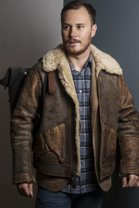 Ein Mann trägt eine braune Lammfelljacke und ein kariertes Hemd und trägt einen Rucksack über der Schulter. Er hat kurze Haare und einen Bart und steht vor einem neutralen Hintergrund.