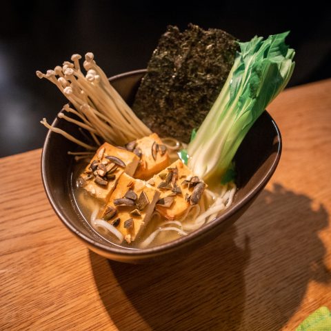 Eine Schüssel Ramen mit Tofu, Enoki-Pilzen, Seetang, Gemüse und einer würzigen Brühe steht auf einem Holztisch. Der Tofu ist mit geschnittenen Pilzen garniert. Die Atmosphäre ist gemütlich und warm.