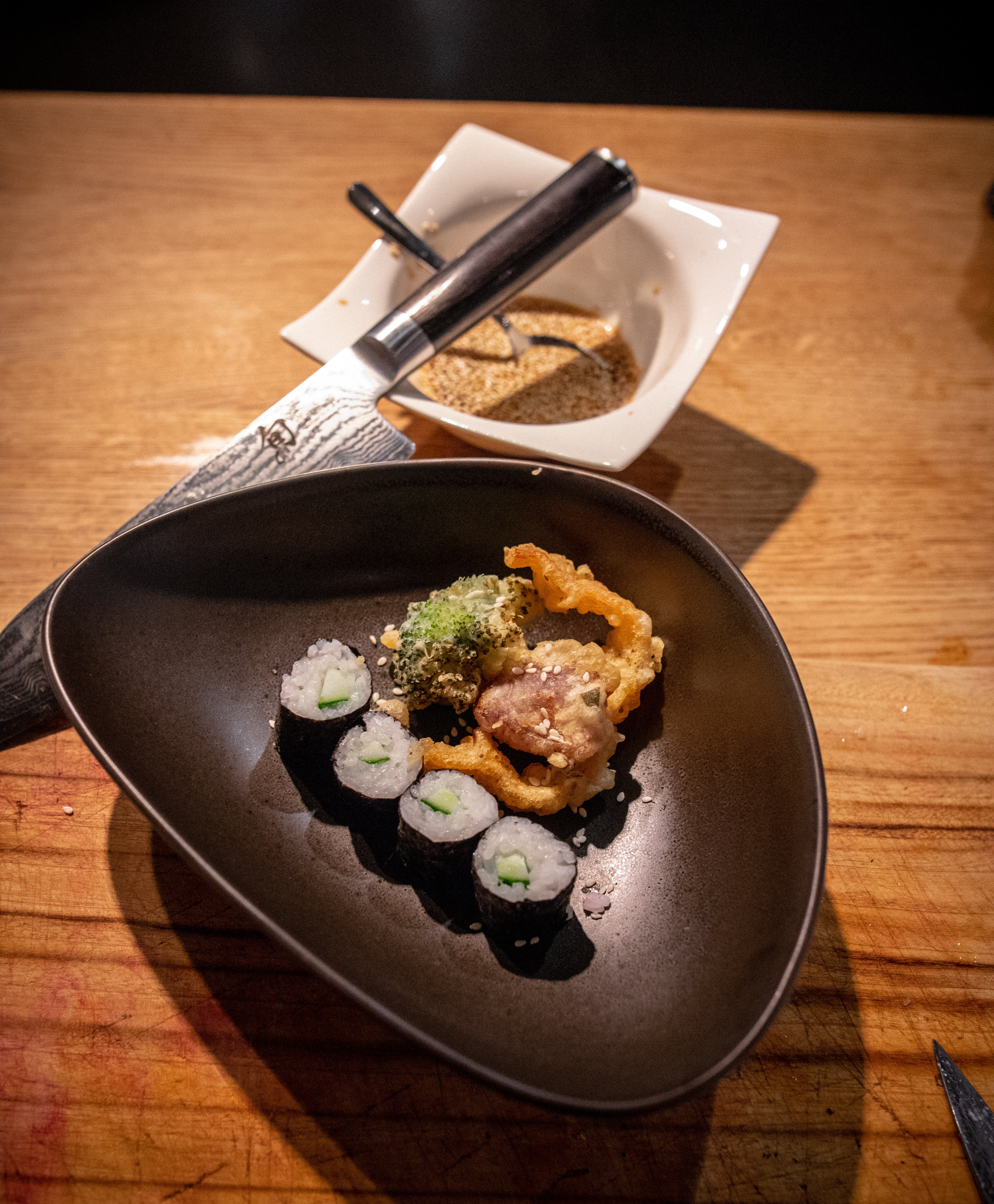 Ein dreieckiger dunkler Teller mit Sushirollen und einem Stück Tempura steht auf einer Holzoberfläche. Im Hintergrund sind eine weiße quadratische Schale mit Dip-Sauce und ein Metallmesser zu sehen.
