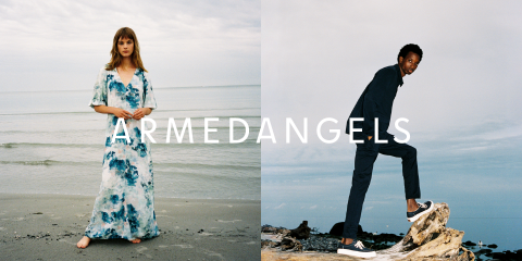 Eine Frau in einem blau-weißen Blumenkleid steht an einem Strand und ein Mann in einem dunklen Outfit posiert auf einem Felsen am Meer. Der Text „ARMEDANGELS“ ist in der Mitte des Bildes eingeblendet.