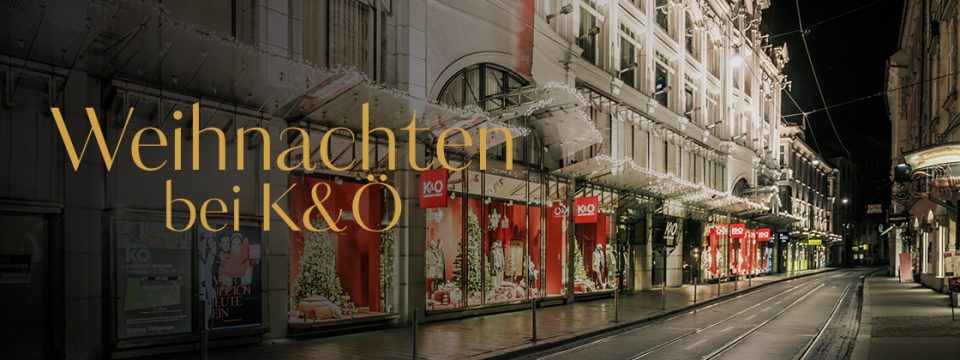 Eine schwach beleuchtete Straße mit Weihnachtsdekorationen und leuchtenden Lichtern vor einem K&O-Laden. Der Text „Weihnachten bei K&O“ ist auf dem Bild deutlich zu sehen.