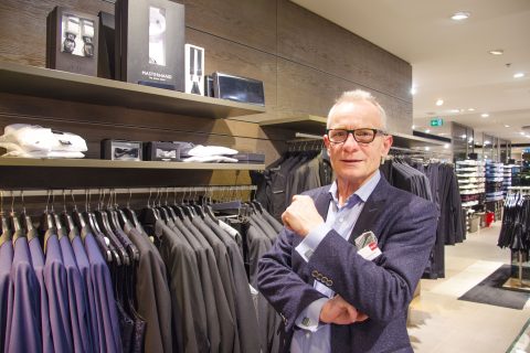 Ein älterer Mann mit Brille und einem marineblauen Anzug steht selbstbewusst in einem Bekleidungsgeschäft. Anzüge und Oberhemden sind auf Ständern um ihn herum ausgestellt, im Hintergrund sind auf Regalen Accessoires zu sehen.