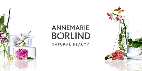 Glasbehälter mit Blumen und Kräutern drumherum, die natürliche Elemente in Szene setzen. In der Mitte steht „ANNEMARIE BÖRLIND NATURAL BEAUTY“. Der Hintergrund ist weiß.