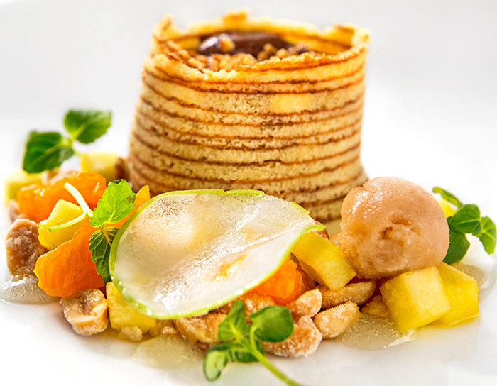 Ein Gourmet-Dessert bestehend aus einem mehrschichtigen Gebäckturm, garniert mit Birnenscheiben, Zitrussegmenten und einer Kugel Sorbet. Rundherum sind frische grüne Kräuter auf einem weißen Teller platziert.