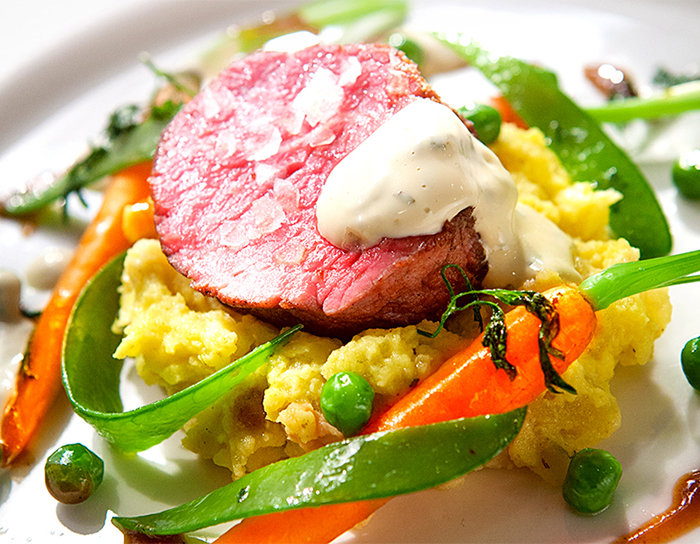 Ein Gourmetgericht mit einer Scheibe blutigem Rindfleisch, garniert mit cremiger weißer Soße, serviert auf einem Bett aus Kartoffelpüree. Dazu gibt es frisches Gemüse wie Zuckerschoten, Karotten und grüne Erbsen, garniert mit frischen Kräutern.
