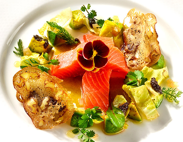 Ein Gourmetgericht mit rohen Lachsscheiben, blumenförmig angeordnet, mit einer essbaren Blume garniert, umgeben von Gemüse, Nüssen und Soße, serviert auf einem weißen Teller.