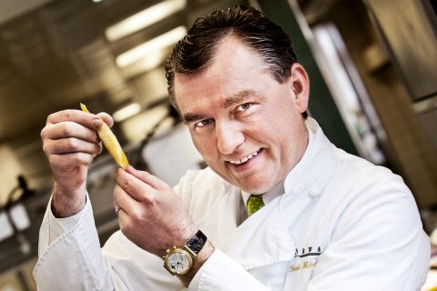 Ein Koch in weißem Kittel und grüner Krawatte hält lächelnd ein Stück Pasta in einer professionellen Küche. Er trägt eine Armbanduhr und im Hintergrund sind Küchengeräte zu sehen.