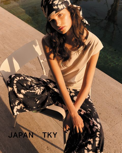 Eine Frau in weißem Top und geblümter Hose sitzt mit verschränkten Armen auf einem Stuhl im Freien. Sie trägt ein passendes geblümtes Kopftuch. Unten steht der Text „JAPAN TKY“. Die Szene ist in warmes Sonnenlicht getaucht.