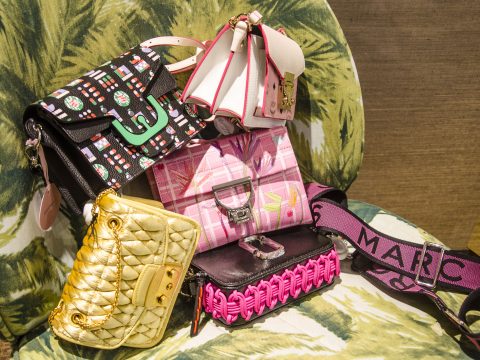 Eine Ausstellung verschiedener Designerhandtaschen in unterschiedlichen Farben und Stilen, darunter eine rosa gewebte Clutch, eine goldene Stepptasche, eine gemusterte schwarze Tasche und andere. Sie sind auf einem Stuhl mit einem Stoff mit tropischem Blattmuster angeordnet.
