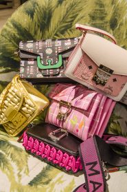 Eine Kollektion farbenfroher Designerhandtaschen auf einem grün gemusterten Hintergrund. Die Taschen variieren im Stil und sind mit Designs wie Karomuster, Metallic-Gold und Stoff mit Stickereien erhältlich. Jede Tasche weist einzigartige Details und Verschlüsse auf.
