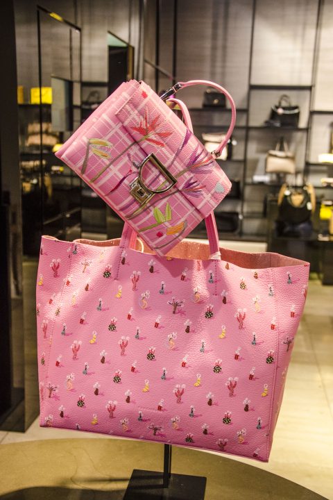 Rosa Handtaschen mit bunten Mustern werden in einem Geschäft ausgestellt. Die größere Tragetasche weist kleine Blumenmuster auf, während die kleinere Handtasche ein Karomuster mit einer Metallschnalle aufweist. Im Hintergrund sind auf Regalen weitere Taschen zu sehen.