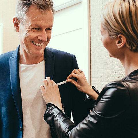 Ein lächelnder Mann in einem blauen Anzug wird von einer Frau mit kurzen Haaren und einer Lederjacke zurechtgerückt. Sie rückt das Revers zurecht, während er zufrieden aussieht. Sie befinden sich in einem gut beleuchteten Raum mit neutral gefärbten Wänden.