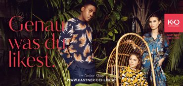 Drei Models in stylischen, gemusterten Outfits stehen und sitzen inmitten üppiger grüner Pflanzen. Der Text lautet „Genau was du likest“. Oben rechts befindet sich ein rotes Logo mit „Kastner & Öhler“. Unten in der Mitte steht die Website-URL.