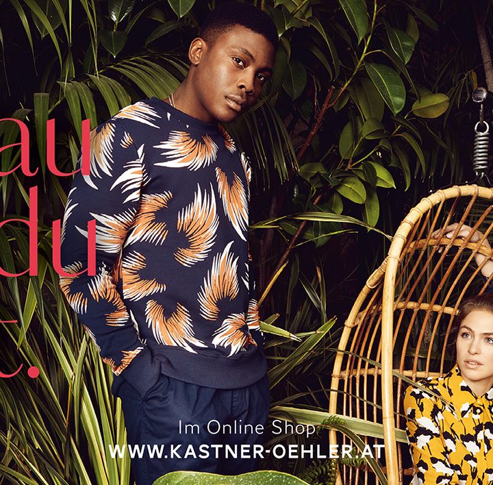 Drei Models in stylischen, gemusterten Outfits stehen und sitzen inmitten üppiger grüner Pflanzen. Der Text lautet „Genau was du likest“. Oben rechts befindet sich ein rotes Logo mit „Kastner & Öhler“. Unten in der Mitte steht die Website-URL.