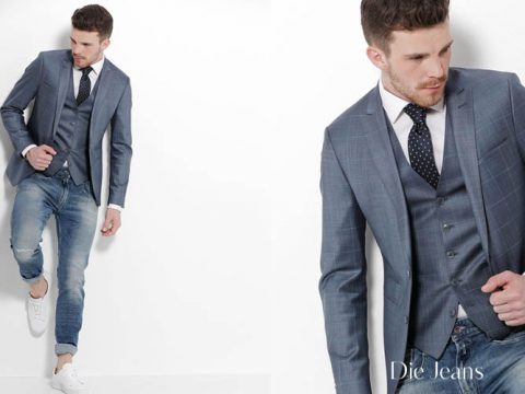 Ein Mann in einem maßgeschneiderten grauen Anzugsjackett und einer Weste, dazu hochgekrempelte Jeans und weiße Turnschuhe. Er trägt ein weißes Hemd und eine gepunktete Krawatte und posiert vor einem weißen Hintergrund. Unten rechts steht der Text „Die Jeans“.