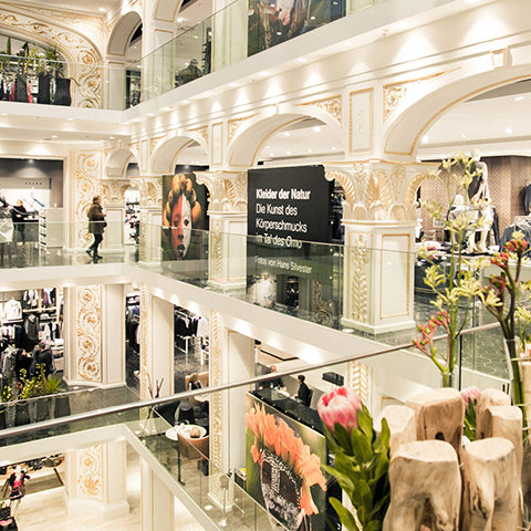 Ein mehrstöckiges Einkaufszentrum mit kunstvoll verzierter Innenausstattung in Weiß und Gold. Käufer stöbern in Geschäften auf verschiedenen Etagen. Große Plakate und Blumenarrangements sind zu sehen. Glasgeländer und dekorative Bögen verleihen der Szenerie Eleganz.