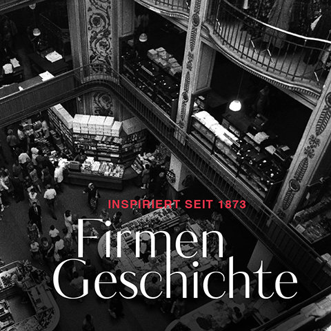 Schwarzweißbild eines geschäftigen mehrstöckigen Kaufhauses mit kunstvollen Dekorationen. Menschen kaufen zwischen Regalen voller Waren ein. In rotem Text steht „Inspiriert seit 1873“ und in großem weißen Text steht „Firmen Geschichte“.