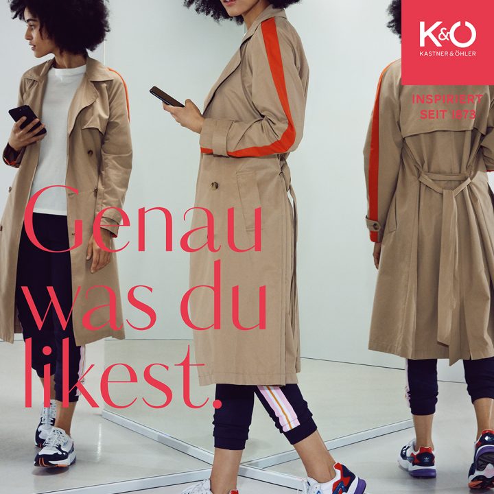 Eine Person mit lockigem Haar, beigem Trenchcoat und Turnschuhen schaut in einem modernen Raum mit Spiegeln auf ihr Handy. Der Text lautet „Genau was du likest“. Oben rechts ist ein rotes Quadrat mit „Kastner & Öhler“ zu sehen.
