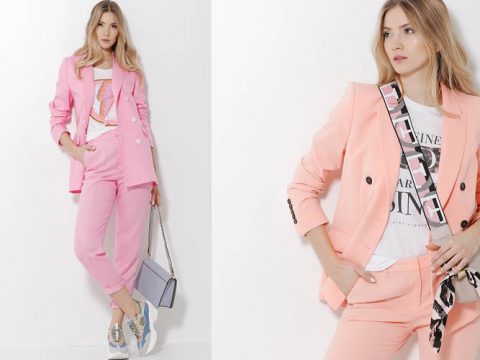 Eine Frau präsentiert ein stylisches Outfit bestehend aus einem hellrosa Blazer und einer passenden Hose. Dazu kombiniert sie ein bedrucktes T-Shirt, Sneakers und eine Umhängetasche. Die Kulisse ist ein schlichter, heller Hintergrund.