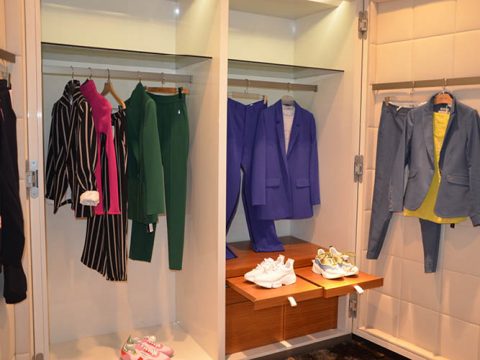 Ein Schrank, in dem verschiedene Damenkostüme in unterschiedlichen Farben ausgestellt sind: schwarz-weiß, grün, lila und blau gestreift. Außerdem stehen zwei Paar Turnschuhe auf einem Holzregal.