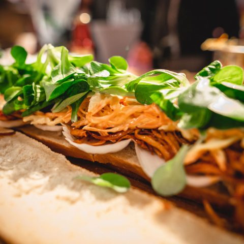 Nahaufnahme eines belegten Sandwichs mit geraspelten Karotten, Gemüse und Zwiebelscheiben auf einer Brotscheibe. Der Fokus liegt auf den frischen Zutaten, der Hintergrund ist unscharf.