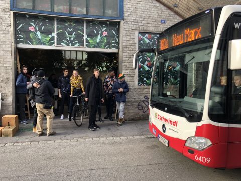 Eine Gruppe von Menschen steht auf einer Straße neben einem Bus mit der Aufschrift „902 New Markt“. Einige halten Fahrräder, andere unterhalten sich oder warten. Im Hintergrund des Gebäudes ist ein Wandgemälde mit großen Blättern und roten Blumen zu sehen. Die Szene wirkt lässig und urban.