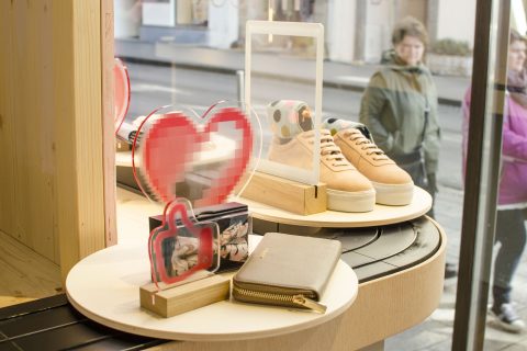 Ein Schaufenster mit beigen Turnschuhen und einer goldenen Geldbörse auf runden Holzregalen. Rote, pixelige Herzen und Daumen-hoch-Schilder sind ebenfalls zu sehen. Vor dem Fenster laufen Menschen auf dem Bürgersteig vorbei.