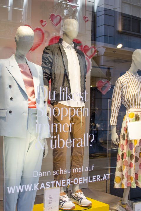 In einem Schaufenster präsentieren Schaufensterpuppen modische Kleidung, darunter einen hellblauen Anzug, eine lässige Jacke mit beiger Hose und einen Rock mit Tupfenmuster. Im Glas sind eine Werbebotschaft auf Deutsch und ein Website-Link zu sehen.