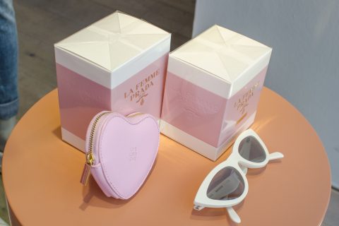 Auf einem pfirsichfarbenen Tisch stehen zwei rosa-weiße Parfümschachteln mit der Aufschrift „La Femme Prada“. Daneben liegen weiße Cat-Eye-Sonnenbrillen und ein kleiner rosa herzförmiger Beutel mit der Aufschrift „LOVE LOVE LOVE“.