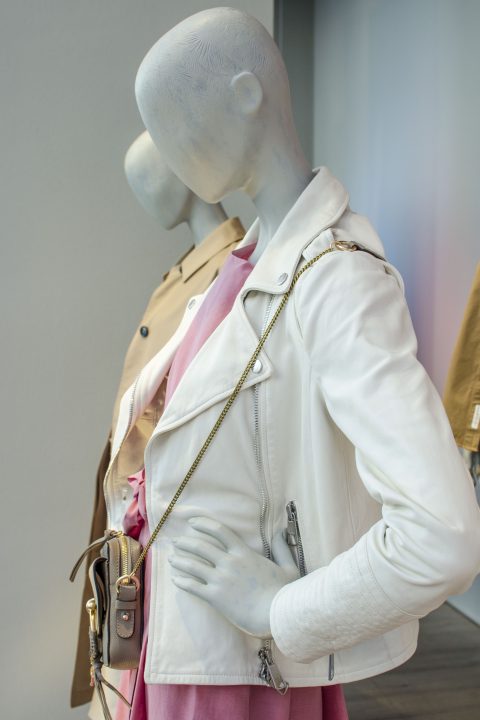 Schaufensterpuppen präsentieren modische Outfits. Die vordere Schaufensterpuppe trägt eine weiße Lederjacke, ein rosa Kleid und eine Umhängetasche. Dahinter trägt eine andere Schaufensterpuppe einen beigen Trenchcoat.