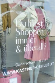Schaufensterdekoration mit Schaufensterpuppen in legerer Kleidung. Eine Schaufensterpuppe trägt eine braune Jacke und Jeans, die andere ein rosa Kleid und eine weiße Jacke. Der Text auf dem Glas lautet: „Du magst Shoppen immer & überall? Dann schau mal auf www.kastner-oehler.at“.
